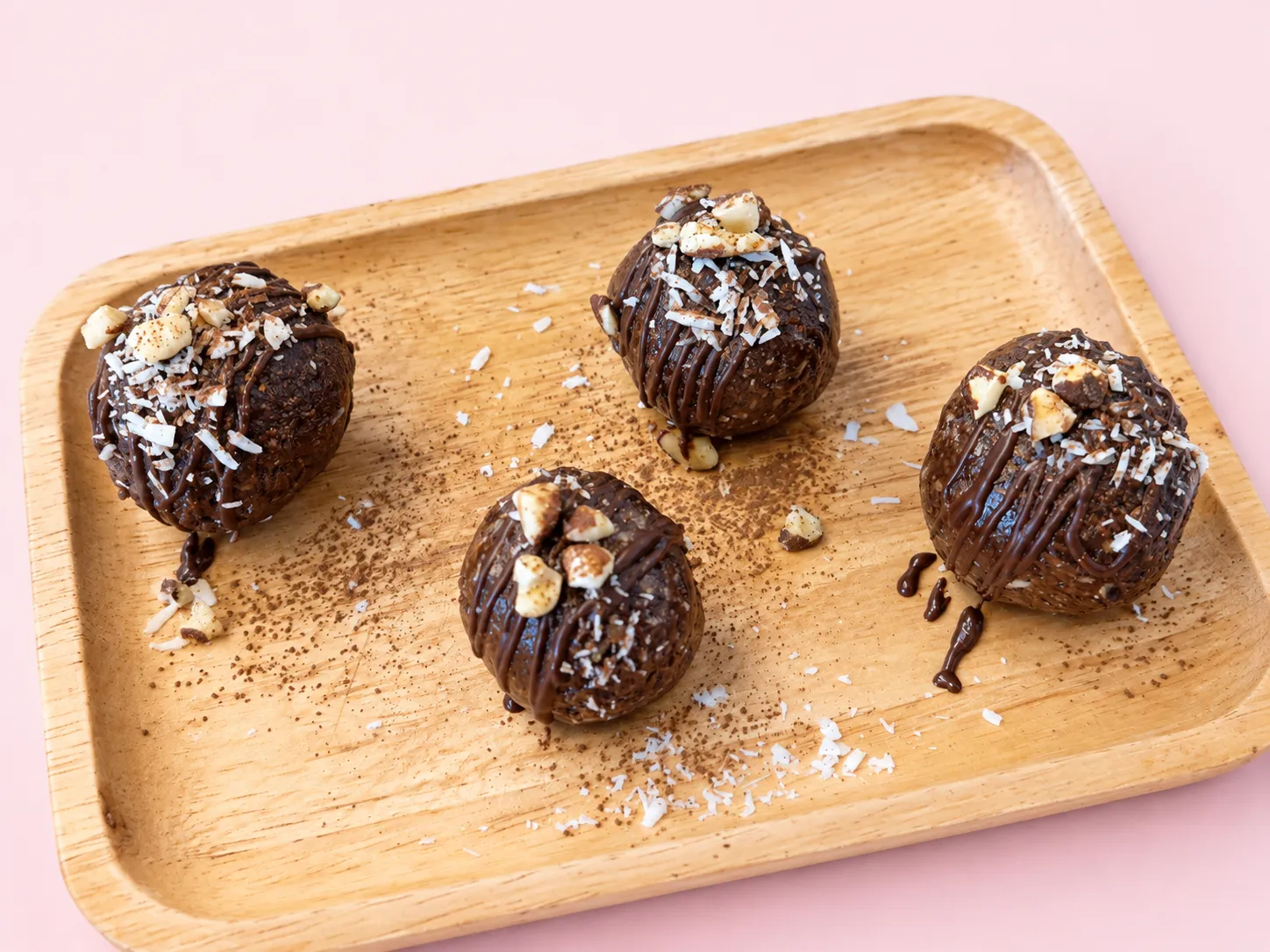 Four chocolate pecan energy bites with a chocolate drizzle, chopped nuts and coconut on a wooden tray
